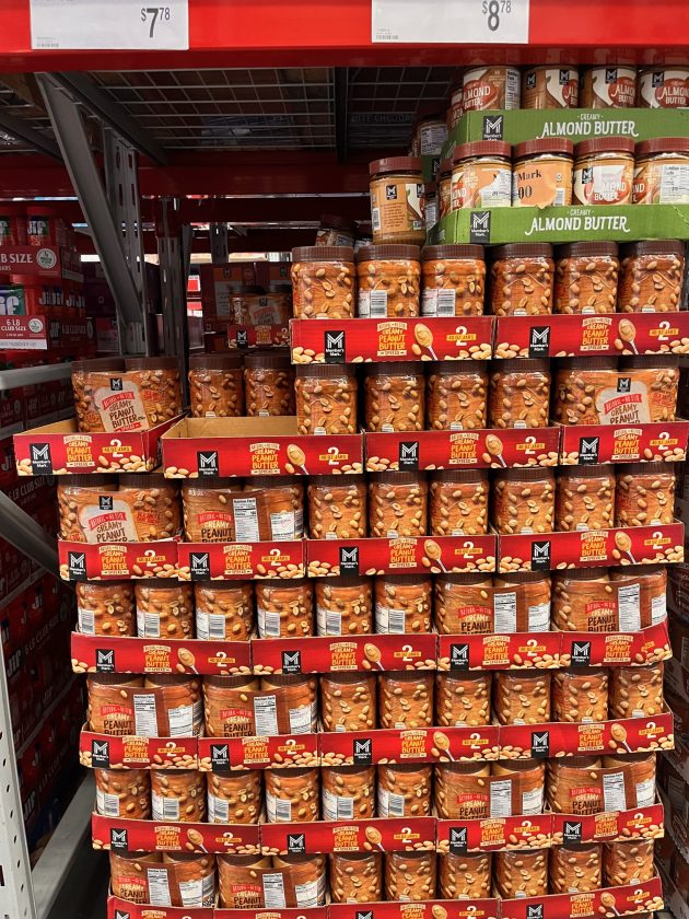 Shelf stacked with jars labeled "CREAMY PEANUT BUTTER" and containers labeled "ALMOND BUTTER" arranged in bulk packaging.