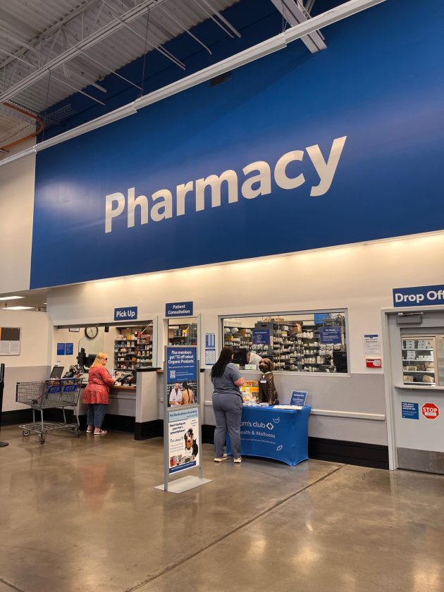 Pharmacy counter area with a large blue sign reading "Pharmacy" and customers standing at pickup and consultation windows.