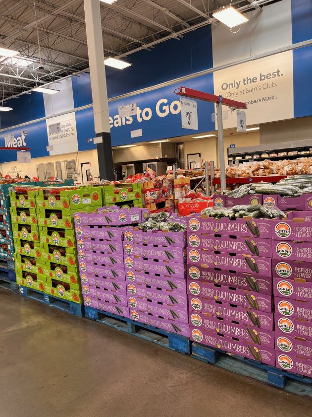 Repeated view of produce pallets filled with boxed cucumbers and other fresh items in a wide warehouse style grocery aisle.