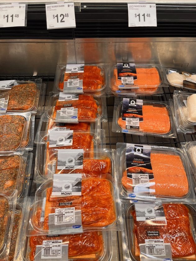 Refrigerated case displaying packaged salmon labeled "Gently Smoked Atlantic Salmon" and other varieties with price signs above showing "$11.48" and "$12.48."
