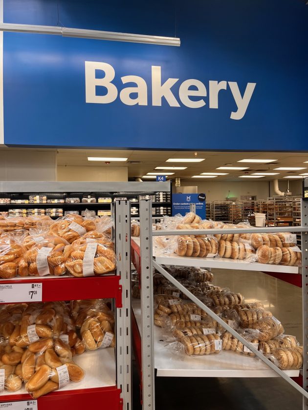 Bakery section in Sam's Club with a blue sign reading "Bakery" above shelves filled with packaged bagels and bread in clear plastic bags.