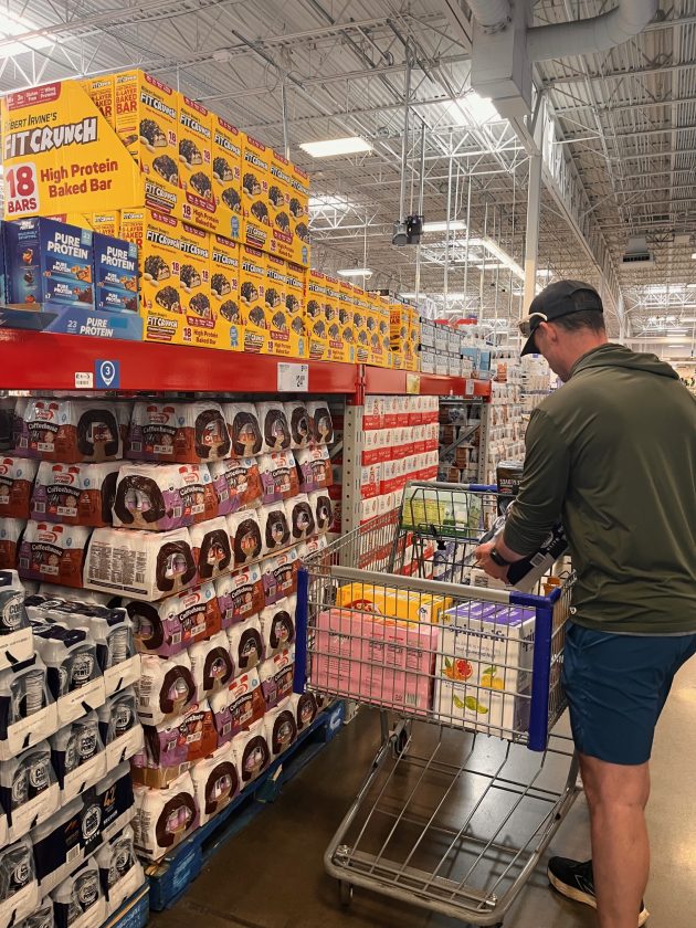 Man pushing a shopping cart through a Sam's Club aisle stacked with bulk snacks and drinks on tall shelves.