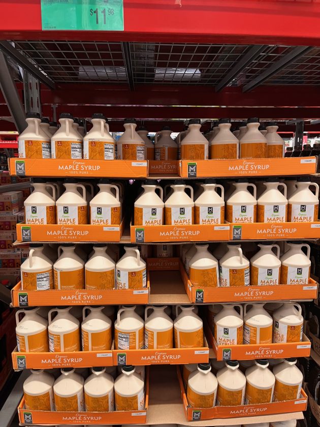 Shelf display of large jugs labeled "MAPLE SYRUP 100% PURE" arranged in rows on a warehouse rack.