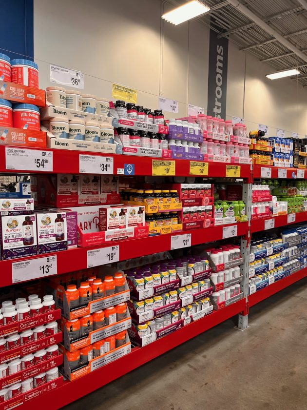 Store aisle stocked with shelves of vitamins and supplements including bottles labeled "Collagen Peptides" and "Bloom" with multiple price tags along the shelves.