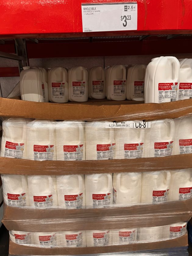 Pallet stacked with gallon jugs labeled "Vitamin D Milk" with a price sign above reading "$3.23" on a warehouse shelf.