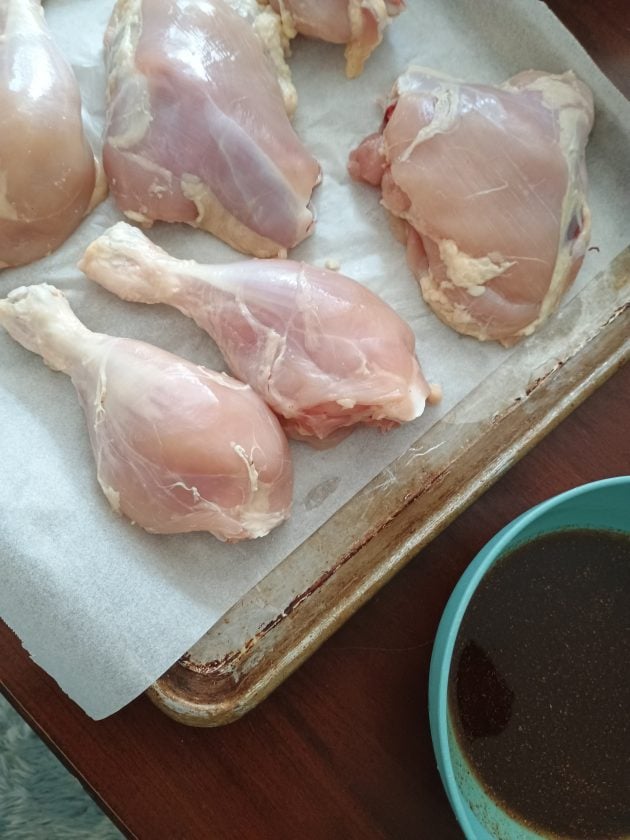 Uncooked chicken leg quarters and drumsticks arranged on a parchment lined baking sheet. A bowl of dark marinade sits beside the pan.