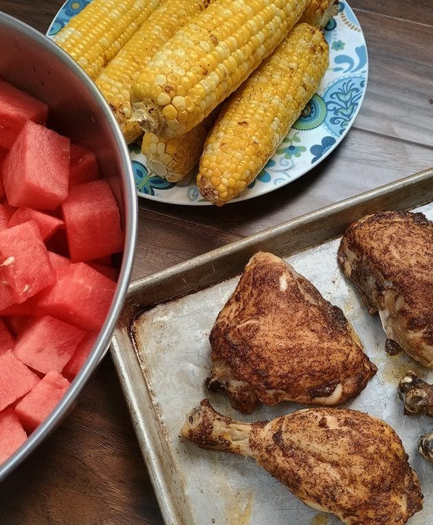 Sheet pan with baked chicken leg quarters and drumsticks beside a bowl of cubed watermelon and a plate of grilled corn on the cob.