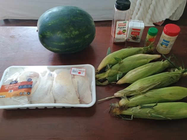Ingredients for a budget friendly family dinner including raw chicken leg quarters, a whole watermelon, fresh corn in husks, and seasonings on a table.