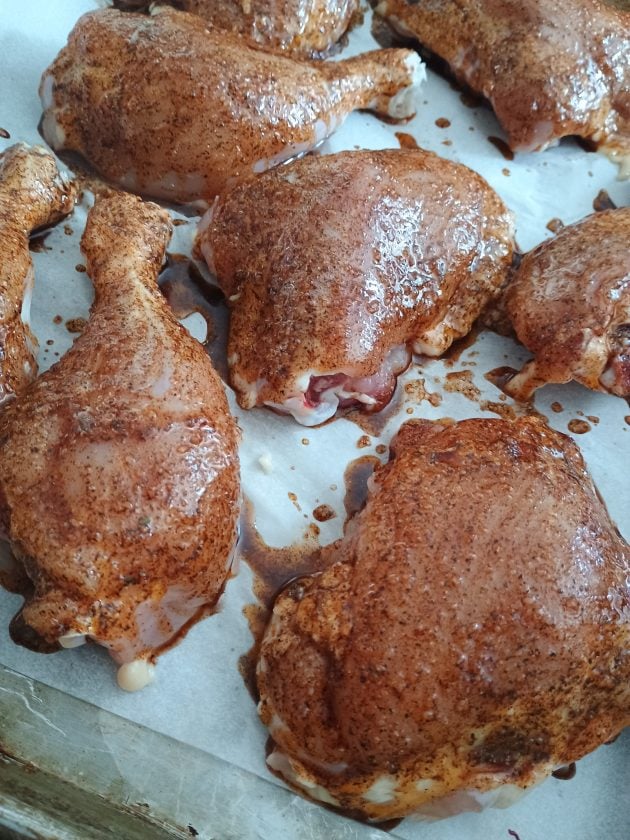 Raw chicken leg quarters and drumsticks coated with seasoning and marinade on a parchment lined baking sheet.