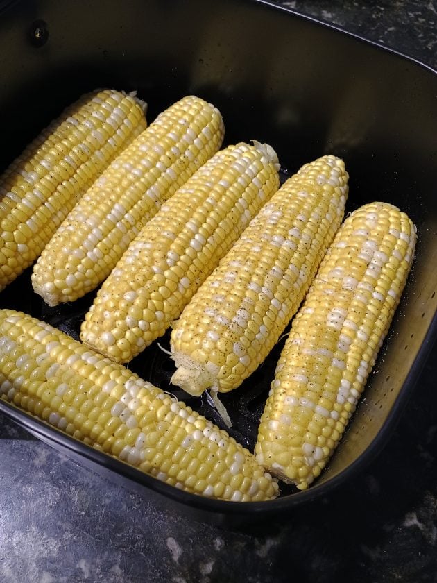 Fresh ears of corn arranged in an air fryer basket before finishing cooking. The corn is coated with butter and black pepper.