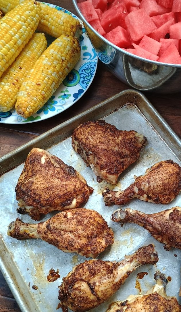 Close up of baked chicken pieces with browned seasoning on a metal baking sheet. A plate of grilled corn and a bowl of watermelon sit in the background as side dishes.
