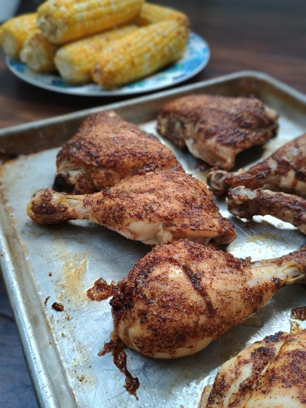 Finished baked chicken with crisp browned skin on a metal baking sheet. Grilled corn on the cob is in the background.
