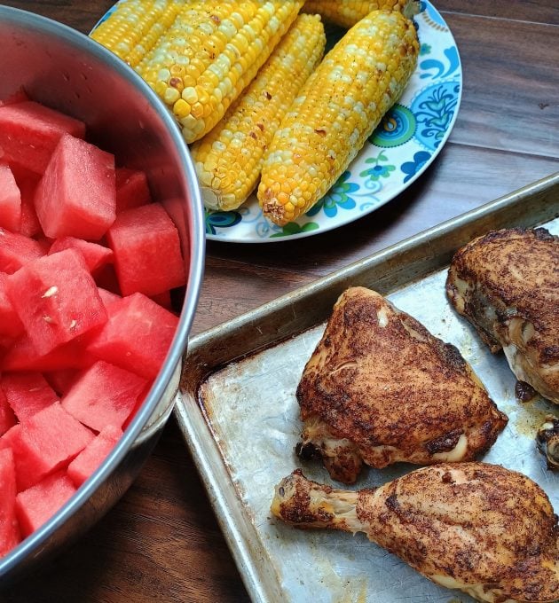 Baked chicken on a sheet pan served with grilled corn on the cob and cubed watermelon. 