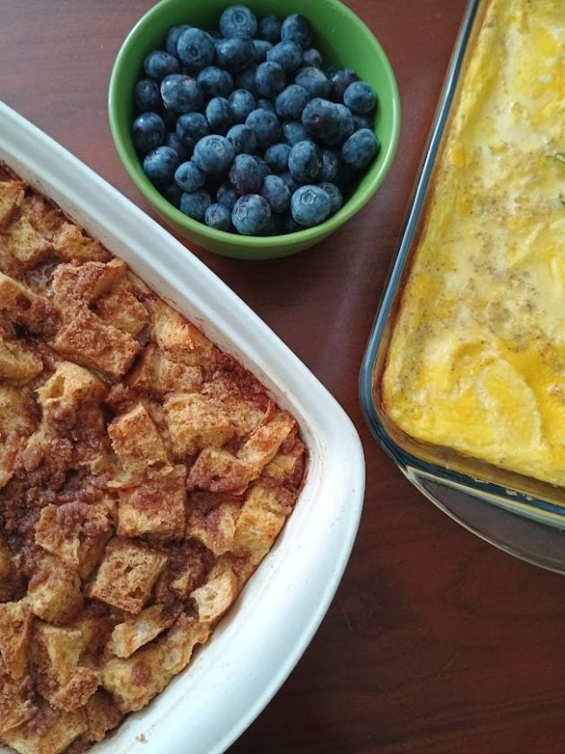Overhead view of a baked French toast casserole, baked scrambled eggs, and fresh blueberries arranged on a wooden table.