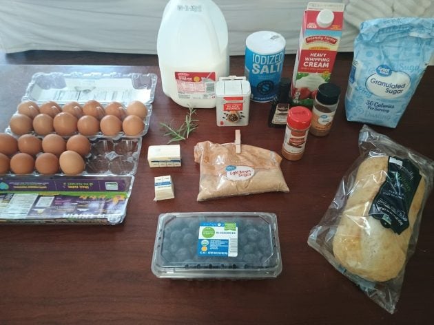Ingredients for French toast casserole and baked scrambled eggs arranged on a table including bread, eggs, milk, blueberries, sugar, brown sugar, cream, butter, cinnamon, vanilla, salt, and fresh herbs.