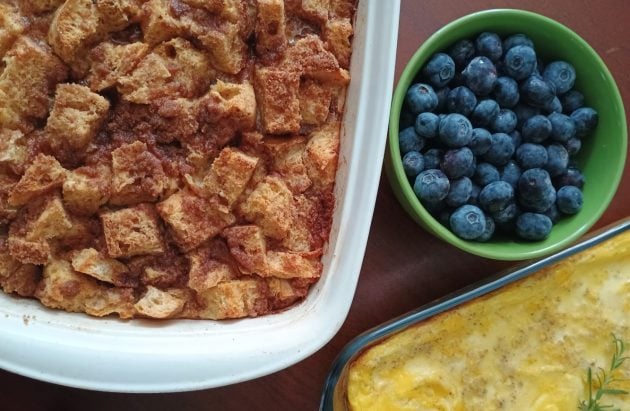 Baked French toast casserole in a white baking dish served alongside baked scrambled eggs and a bowl of fresh blueberries on a wooden table.