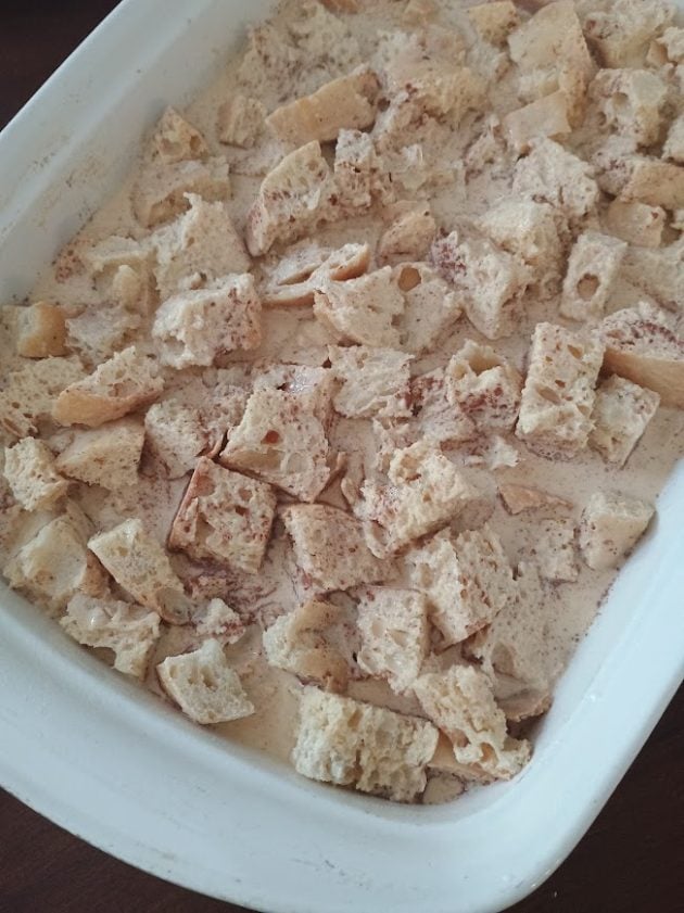 Cubed bread soaking in a cinnamon egg custard mixture inside a white baking dish.