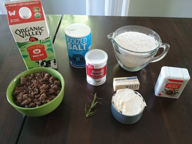 Ingredients for homemade biscuits and gravy arranged on a table including flour, milk, sausage, butter, sour cream, baking powder, salt, pepper, and fresh rosemary.