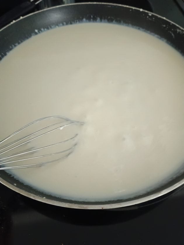 White gravy being whisked in a black skillet on the stovetop while cooking homemade biscuits and gravy.