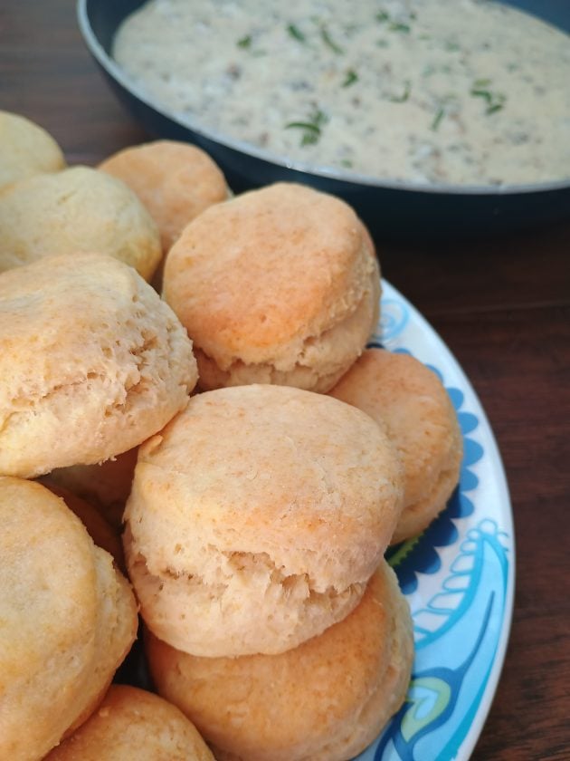 tack of fluffy homemade biscuits on a patterned plate with a skillet of creamy sausage gravy.