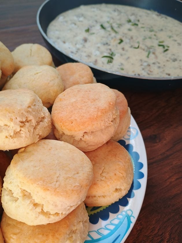 Homemade biscuits stacked on a patterned plate beside a skillet of creamy sausage gravy topped with chopped herbs.