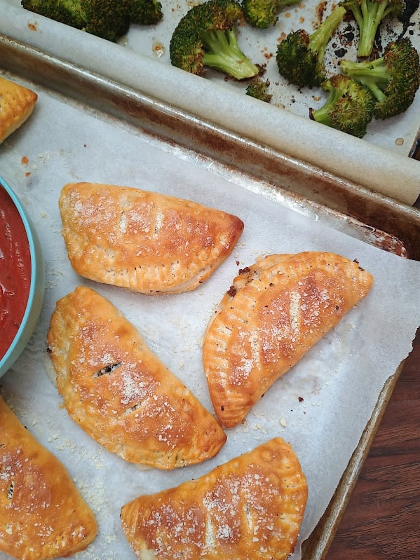 Baked mini calzones with roasted broccoli and a bowl of marinara sauce on the side.
