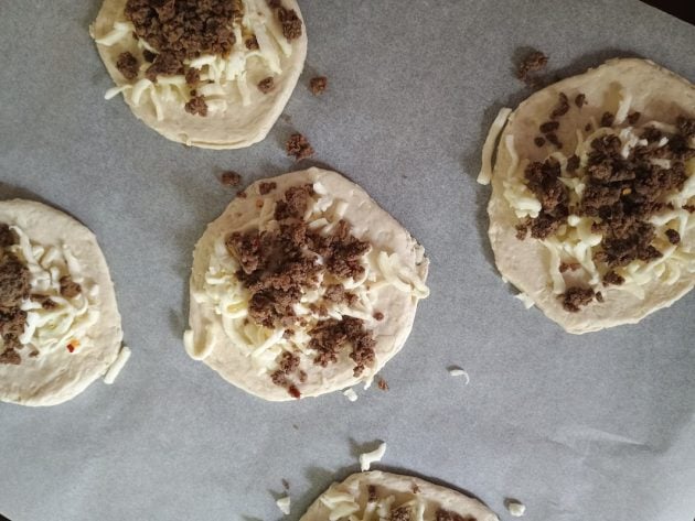 Biscuit dough rounds topped with shredded mozzarella and cooked ground beef for mini calzones.
