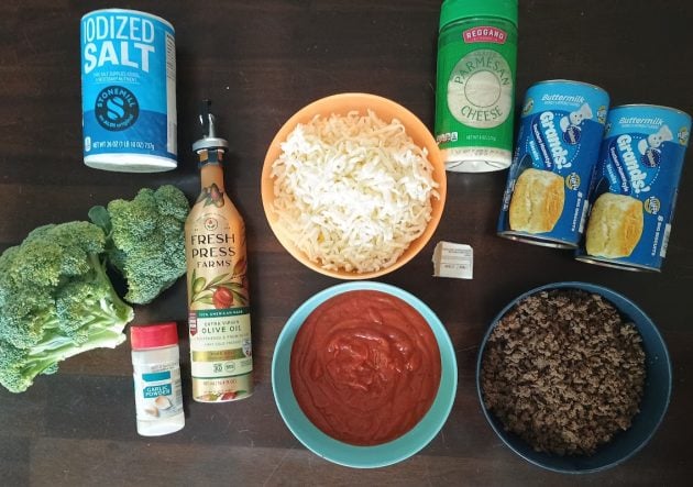 Ingredients for mini calzones arranged on a table including biscuit dough, mozzarella, ground beef, sauce, broccoli, olive oil, garlic powder, salt, and Parmesan cheese.