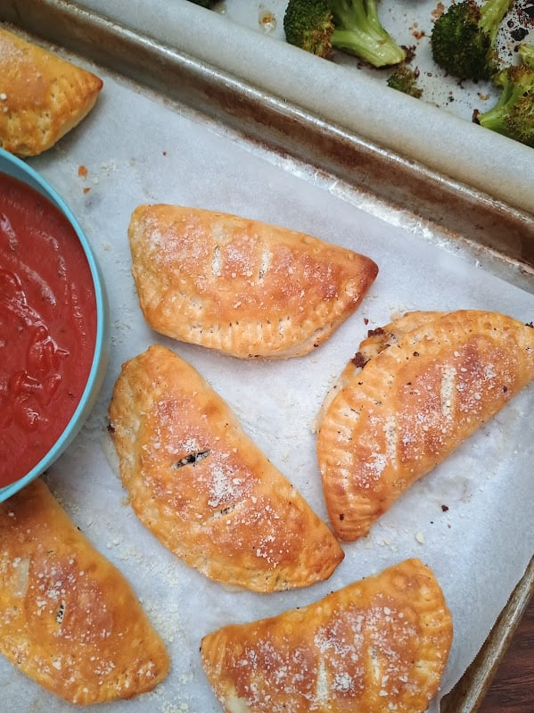 Baked mini calzones with roasted broccoli and a bowl of marinara sauce on the side.