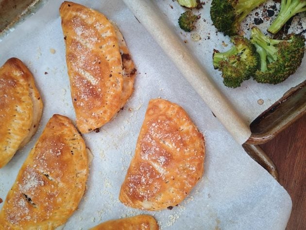 Golden baked mini calzones on parchment paper with roasted broccoli in the background.