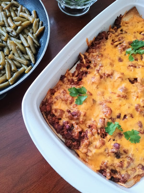 White casserole dish filled with cheesy Mexican lasagna served beside a pan of green beans.