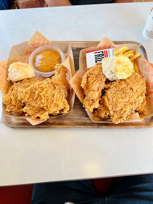 Two trays of crispy fried chicken served with biscuits, fries, and a side of gravy on a restaurant table.
