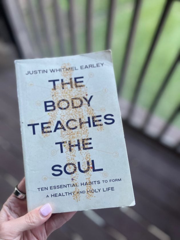 Hand holding the book "The Body Teaches the Soul. Ten Essential Habits to Form a Healthy and Holy Life" by Justin Whitmel Earley in front of an outdoor railing.