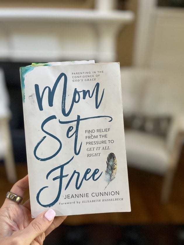 Hand holding the book "Mom Set Free. Find Relief from the Pressure to Get It All Right" by Jeannie Cunnion with a living room in the background.
