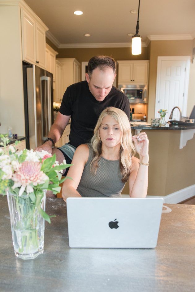 Crystal Paine sits at a kitchen table working on a laptop while her husband, Jesse, leans over her shoulder and looks at the screen with her.