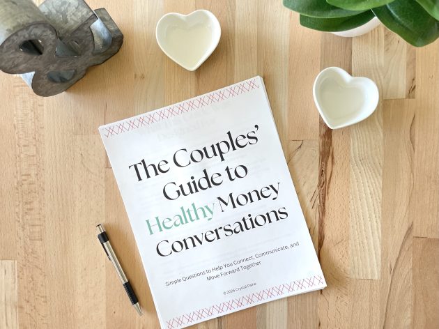A printed workbook titled "The Couples' Guide to Healthy Money Conversations" lies on a wooden table beside a pen and two heart shaped dishes. The cover reads "Simple Questions to Help You Connect. Communicate. and Move Forward Together" and "© 2026 Crystal Paine."