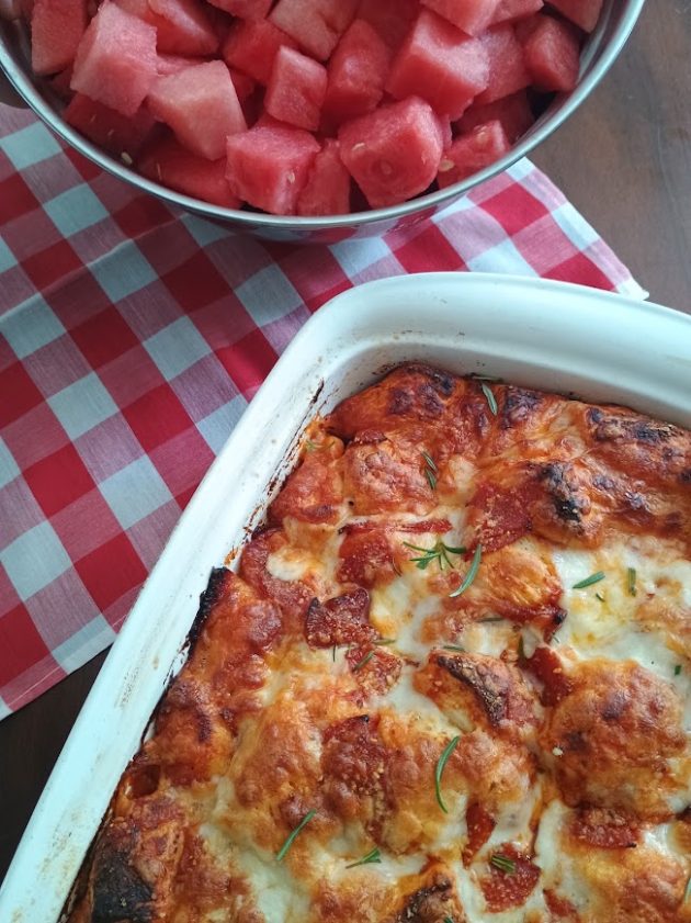 Golden brown bubble pizza topped with melted cheese pepperoni and fresh rosemary. A large bowl of watermelon and a red and white checkered napkin complete the meal.