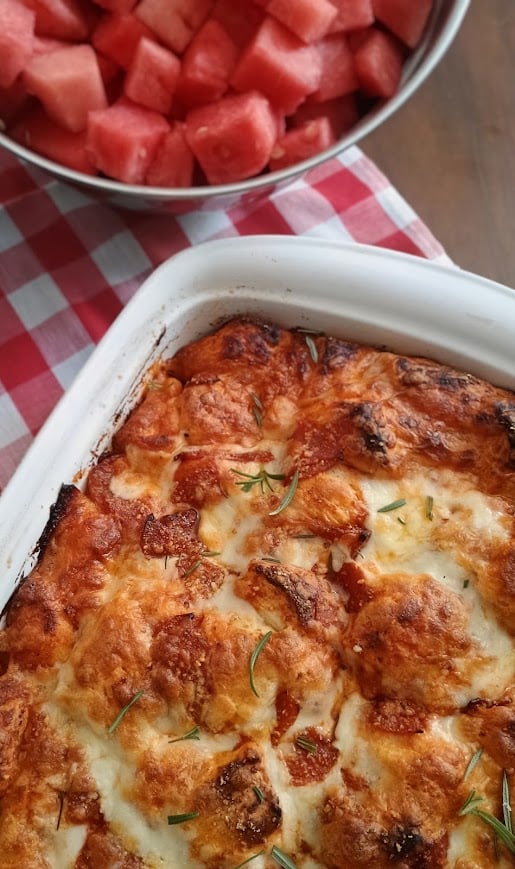 Freshly baked bubble pizza with bubbly browned cheese pepperoni and rosemary in a white casserole dish. Cubed watermelon is served alongside it on a red and white checkered napkin.
