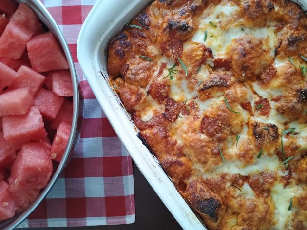 Baked bubble pizza in a white casserole dish with browned melted cheese and pepperoni. A bowl of cubed watermelon sits alongside the pizza.