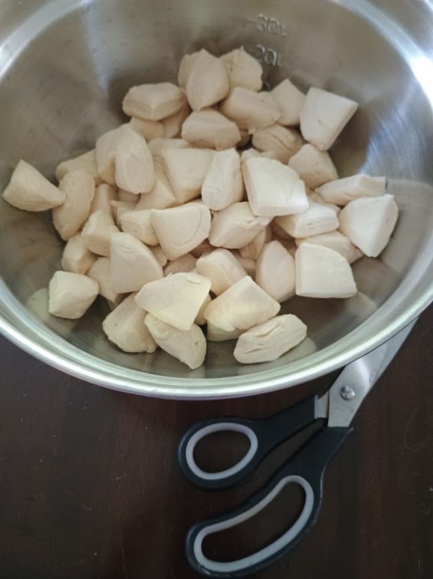 Pieces of refrigerated biscuit dough cut into bite sized chunks in a metal mixing bowl. Kitchen scissors beside the bowl.