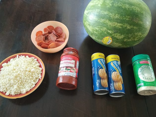 Ingredients for bubble pizza arranged on a table including biscuit dough pepperoni pizza sauce mozzarella Parmesan cheese and a whole watermelon.