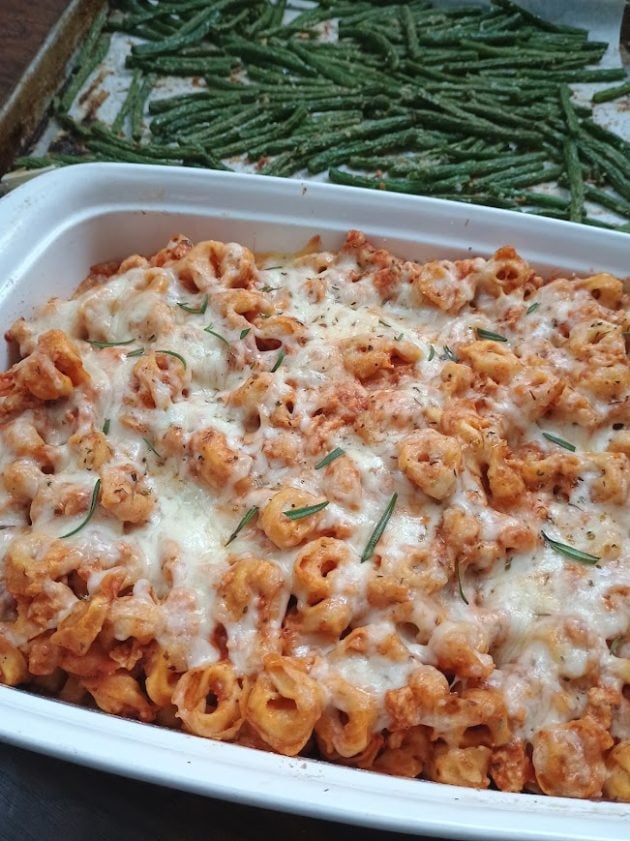 Baked tortellini covered with melted cheese and herbs in a white casserole dish. A tray of roasted green bean fries sits behind it.