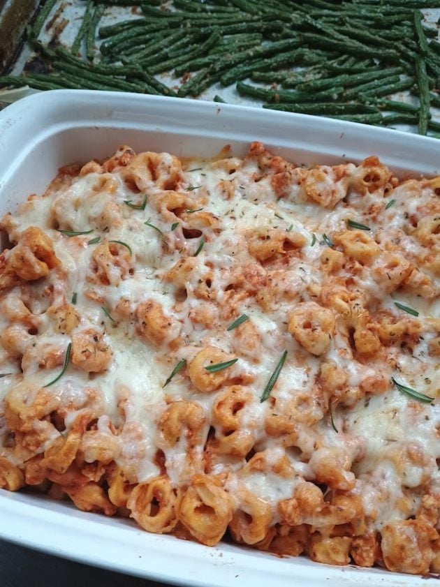 Finished tortellini bake in a white casserole dish with melted golden mozzarella cheese and fresh rosemary scattered over the top. A tray of roasted green beans sits behind the casserole.