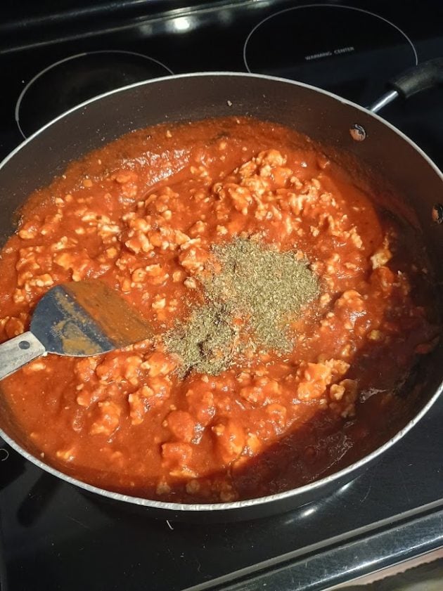 Tomato meat sauce simmering in a large skillet with dried herbs sprinkled in the center and a spatula resting in the sauce.