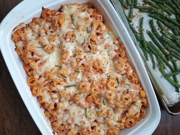 Baked tortellini covered in melted cheese and rosemary in a white casserole dish with a tray of roasted green beans alongside it.