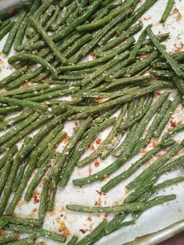 Roasted green bean fries spread across a parchment lined baking sheet with browned seasonings and bits of grated cheese