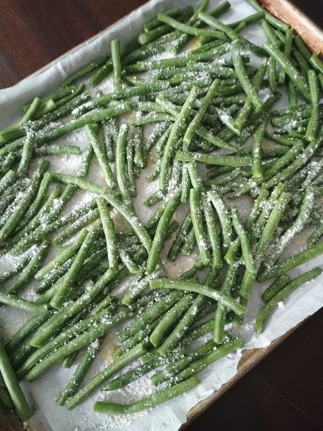 Green bean fries spread across a parchment lined baking sheet before roasting.