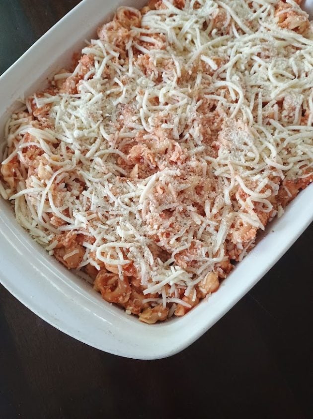 Unbaked tortellini bake in a white casserole dish topped generously with shredded mozzarella cheese and grated Parmesan.