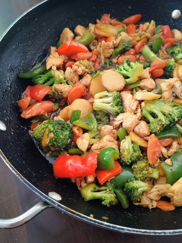 Broccoli, sliced carrots, and red and green bell peppers cooking together in a black skillet, preparing the vegetables for chicken stir fry.