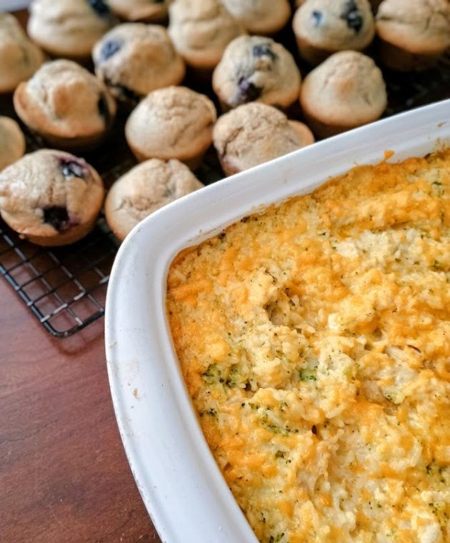 Finished chicken broccoli rice casserole with a golden cheesy topping in a white baking dish. Freshly baked blueberry muffins sit on a cooling rack behind the casserole.